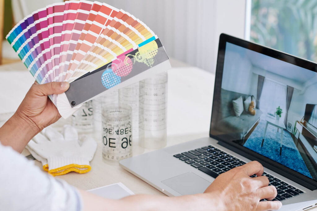 Persona sosteniendo una muestra de colores mientras trabaja en una laptop, revisando un diseño de interiores. En la mesa se observan guantes y frascos con medidas, indicando un proyecto de decoración o renovación.