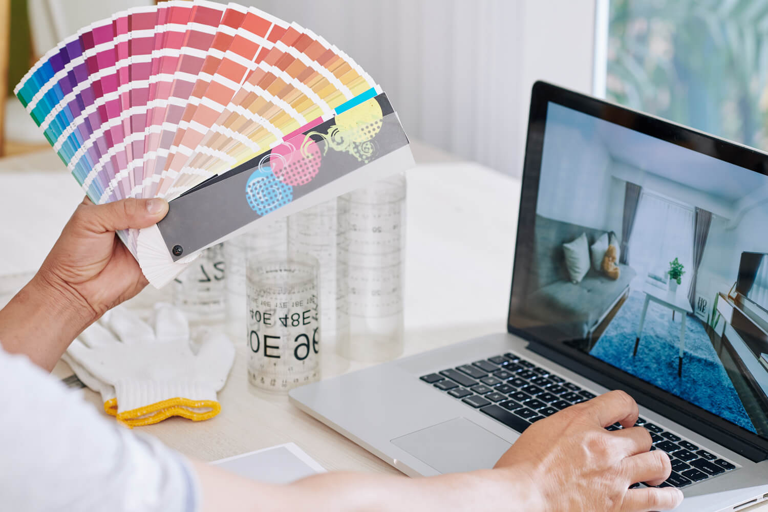 Persona sosteniendo una muestra de colores mientras trabaja en una laptop, revisando un diseño de interiores. En la mesa se observan guantes y frascos con medidas, indicando un proyecto de decoración o renovación.